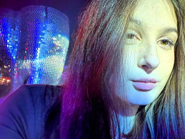 VikVikLove's face with long, slightly messy brown hair, light skin, and pink lips, wearing a black top, illuminated by blue and purple lights. Boobs not visible.