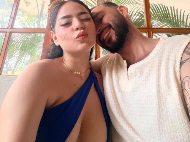 EVAANDWALLE with big tits in a blue dress, arm around bearded dude in white shirt, smiling, brown hair, glasses on head, colorful necklace, tropical window background.