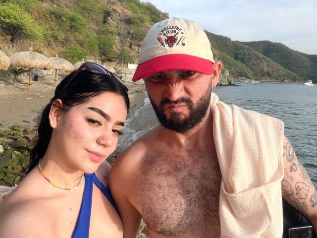 EVAANDWALLE has a cute face with dark hair and blue bikini top, standing next to a hairy, bearded man with a red cap and towel, on a beach.