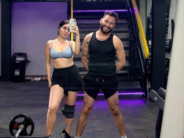 EVAANDWALLE with small tits in a gray sports bra, black skirt, and knee brace, standing next to a bearded guy in a black tank and shorts, smiling in a gym.