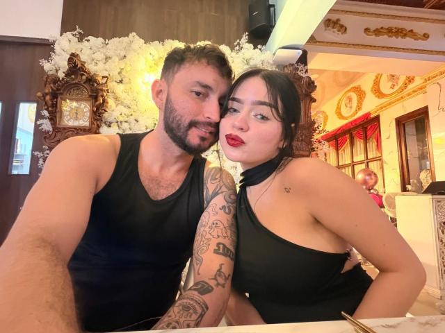 EVAANDWALLE and a tattooed dude with a beard, wearing black tank tops, are in a fancy room with white flowers. She's got red lips and a cute ass.