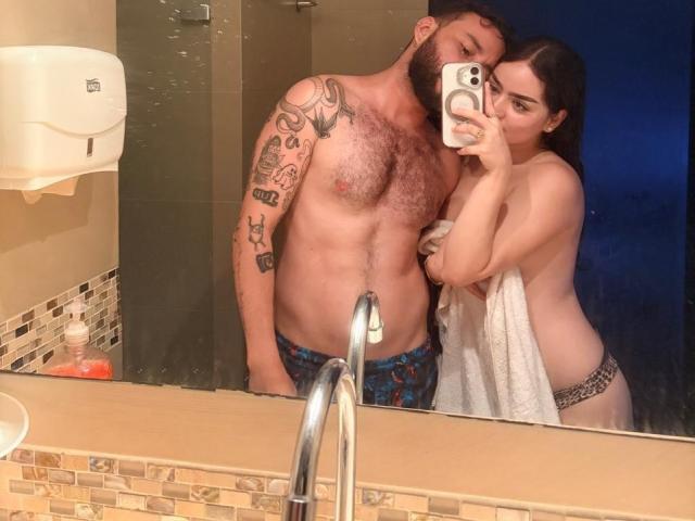 EVAANDWALLE with a beard and tattoos, wearing blue patterned briefs, stands by a mirror as she takes a selfie with her phone, covering her tits with a towel.
