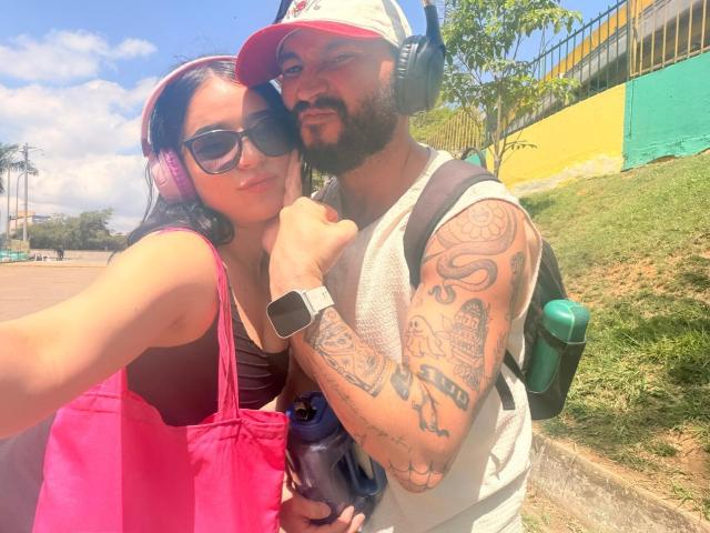 EVAANDWALLE with pink headphones, black sunglasses, pink bag, and brown top, standing next to a bearded man with tattoos, headphones, white tank top, and green backpack.