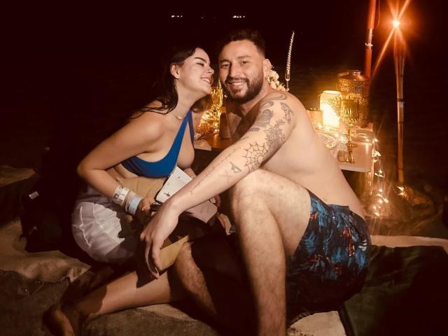EVAANDWALLE with tits in a blue bikini, and WALLE with a beard, tattoos, and blue patterned shorts, sitting on sand, smiling, holding a guitar, under dim light.