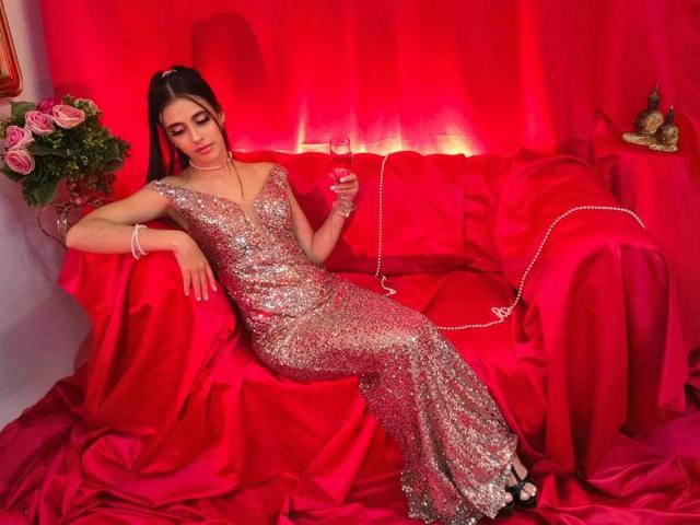 AnnaAlvarez in a shiny, silver sequined dress, reclining on a red satin-covered sofa, holding a glass, with pink roses and a jewel on the right. Red light.