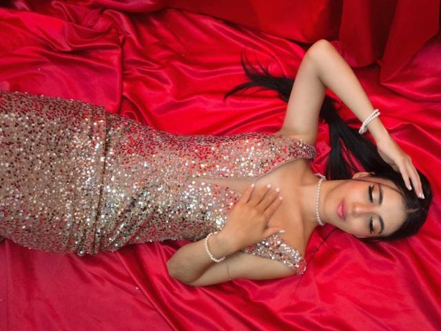 AnnaAlvarez is lying on red silk, wearing a sparkly, sequined dress, with her hand on her chest and the other on her head, eyes closed, black hair spread out.