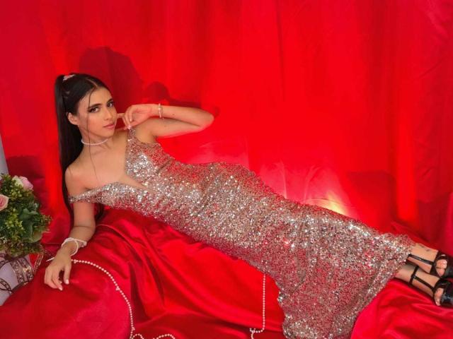 AnnaAlvarez in a sparkly silver dress, lying on red satin, showing her tits, wearing black strappy heels, with a pearl necklace and bracelet, looking sexy.