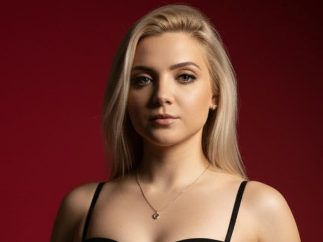 Malvvy has blonde hair, light skin, and is wearing a black strap top. She's looking at the camera with a slight smile, wearing a small necklace.