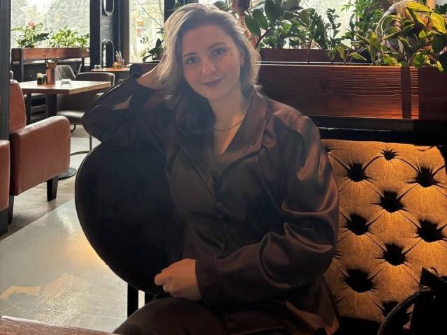 SamuelaBella with big tits, sitting on a tufted, brown, leather couch, wearing a dark brown, shiny, button-up shirt, smiling, hand in hair, in a cozy, plant-filled café.