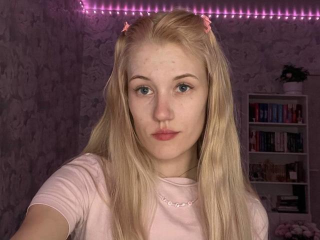 Blonde XuanMaltese with blue eyes, pink hair clips, freckles, and pouty lips, wearing a pink shirt, in a softly lit room with pink LED strip lights.