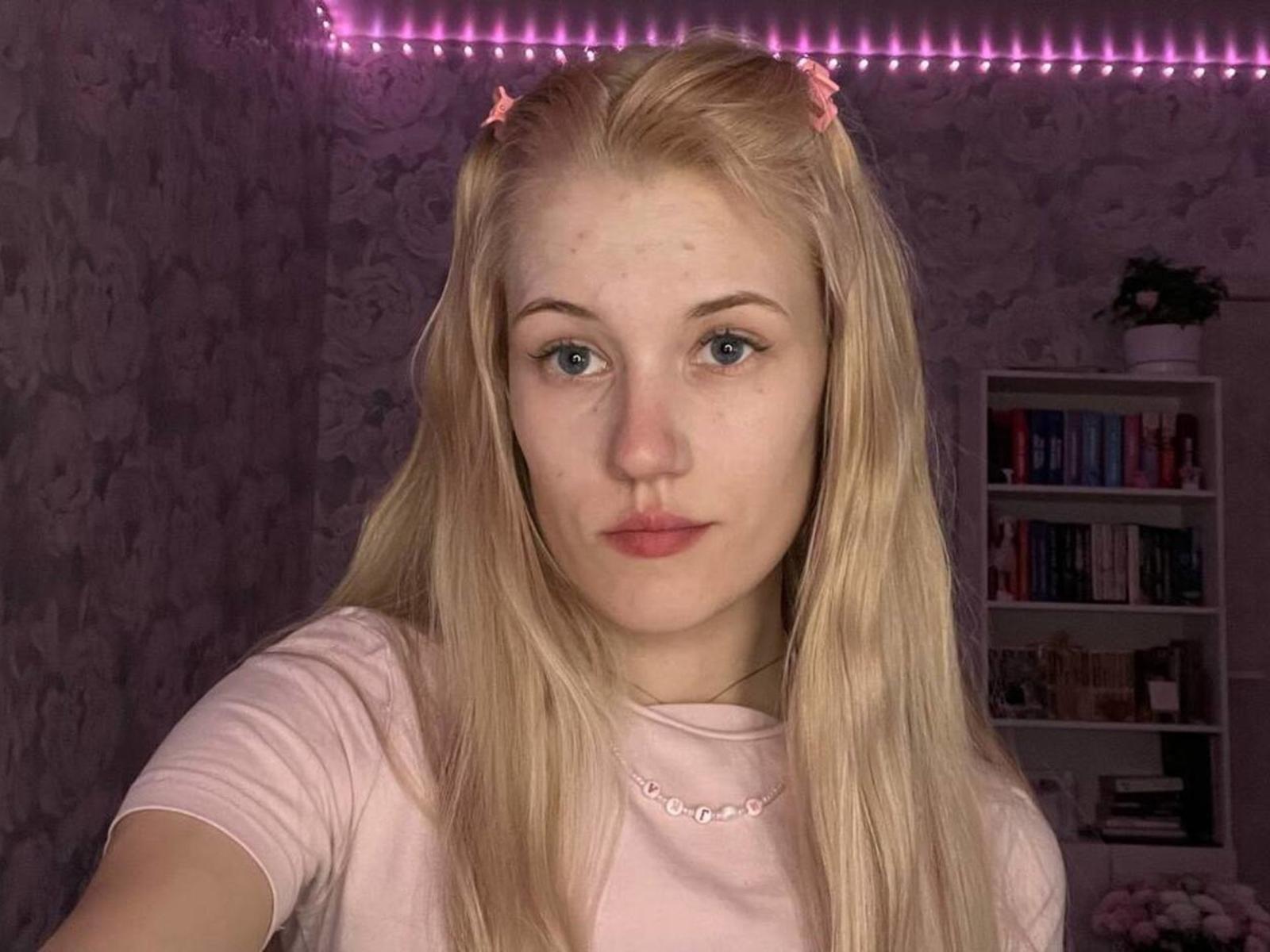 Blonde XuanMaltese with blue eyes, pink hair clips, freckles, and pouty lips, wearing a pink shirt, in a softly lit room with pink LED strip lights.