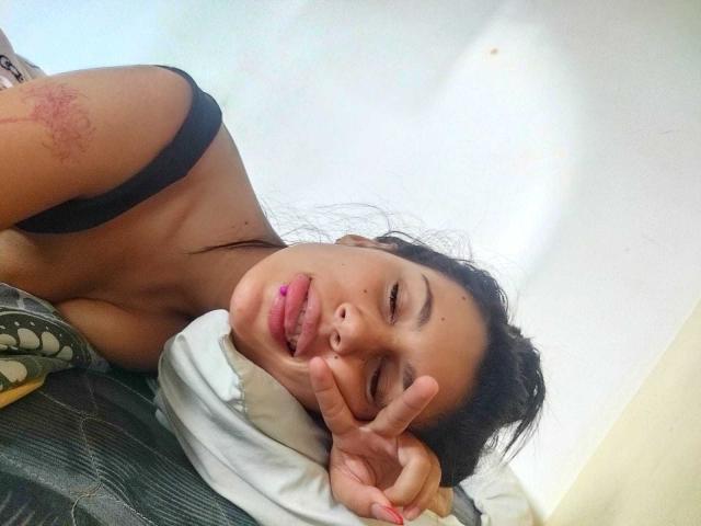 KataleyaMansson with her tongue out, black bra strap visible, red scratch on her arm, making a peace sign, lying on a green bed with a white pillow.