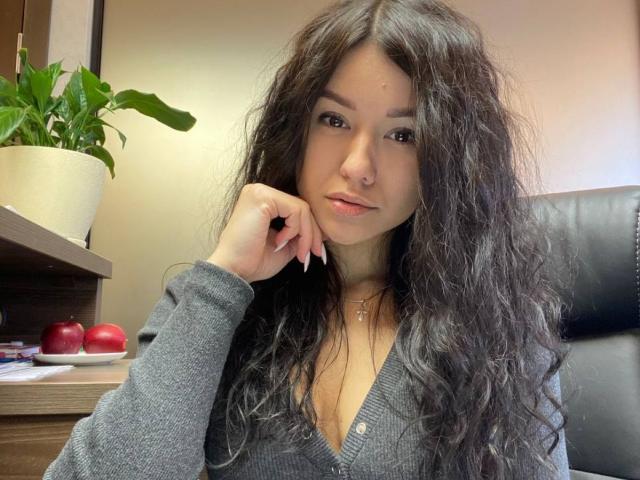 CaramelMills with long, wavy black hair, gray cardigan, and subtle makeup, resting her chin on her hand, showing her small, cute tits and a nose ring.