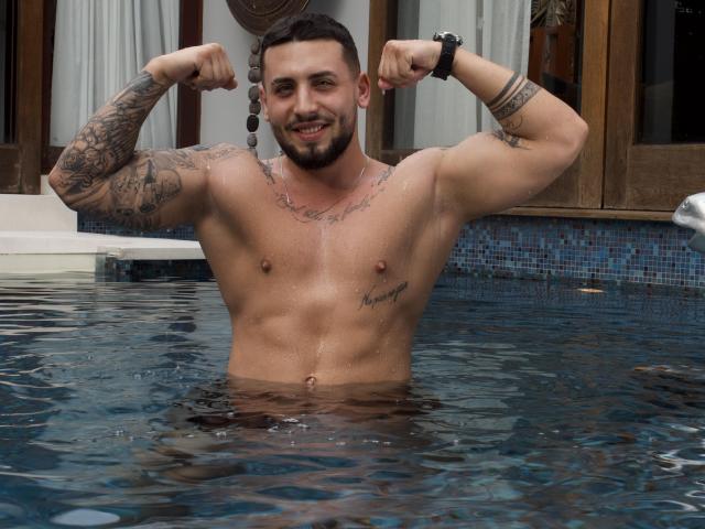GreenDylan, shirtless with tattoos, flexes his muscular arms in a pool, showing off a defined chest and abs, with a confident smile and short beard. Water covers his lower body.