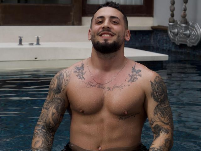 GreenDylan, tattooed, muscular, shirtless, standing in a pool, water droplets on his skin, smiling, short dark hair, beard, chest tattoos, necklace, brown eyes, looking happy.