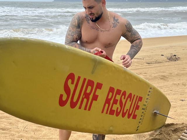 Shirtless GreenDylan with tattoos, holding yellow surf rescue board, looking at red item in hand, standing on sandy beach, waves in background, slight beard, short hair, red board shorts.
