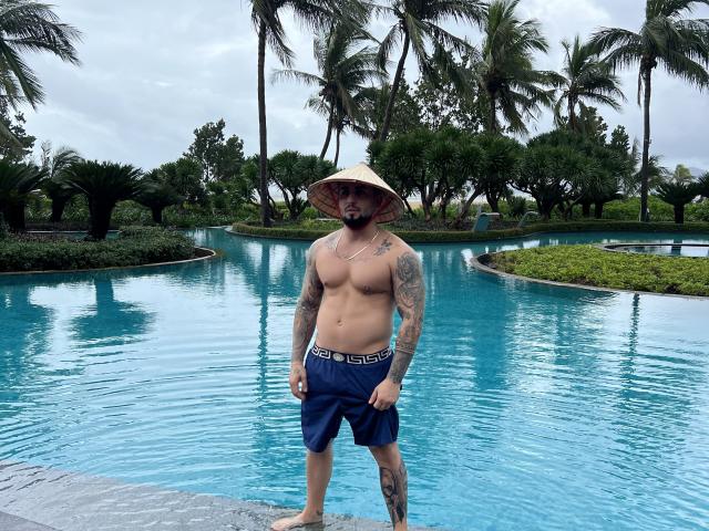 GreenDylan, shirtless with tattoos, wearing blue shorts and a conical hat, standing in a blue-tiled pool, showing a muscular, tattooed, bearded, white-skinned body.