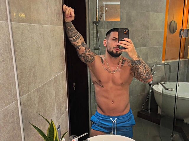 GreenDylan with tattoos, muscular, shirtless, wearing blue shorts, taking a mirror selfie, right arm raised, phone in left hand, beard, pouty expression, in a modern bathroom.