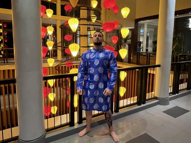 GreenDylan, a bearded dude with tattoos, stands barefoot in a blue dress with white patterns, surrounded by glowing red and yellow paper lanterns, in a hotel hallway.