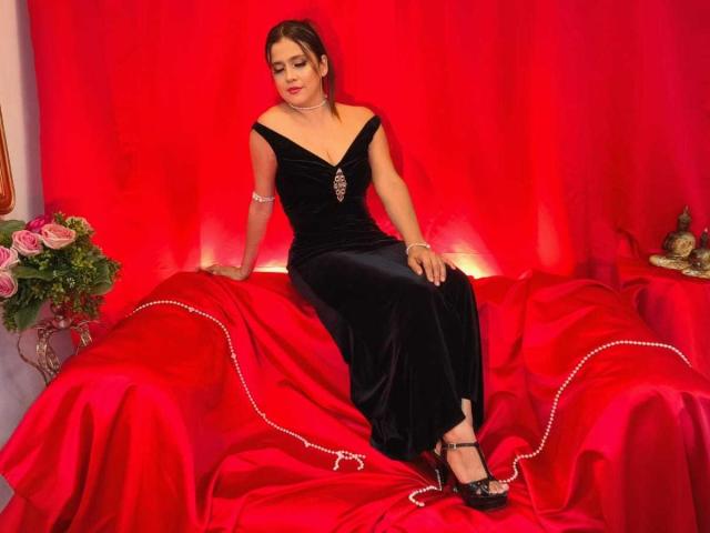 HeidyWells in a black, off-shoulder, velvet dress, sitting on a red, satin-covered couch, with a pearl bracelet, red lips, and black heels, looking down.