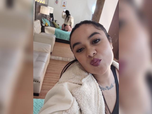 HannahMolly with puffy purple lips, black hair in a ponytail, wearing a white fuzzy jacket, black tank top, and necklace, standing in a bedroom, mirror selfie.