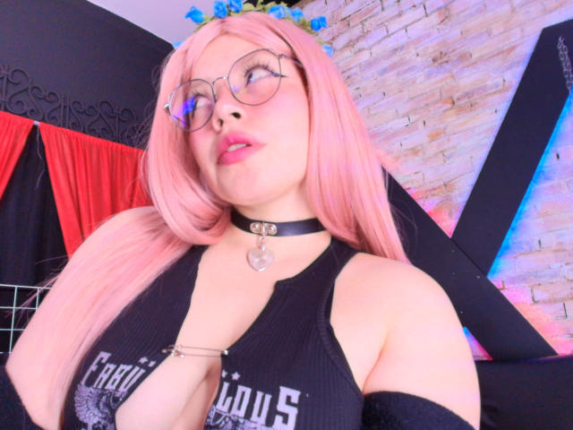 Nyxaria with pink hair, glasses, and blue flower crown, wearing a black choker with heart, and a black top showing her tits, looking up with parted pink lips.