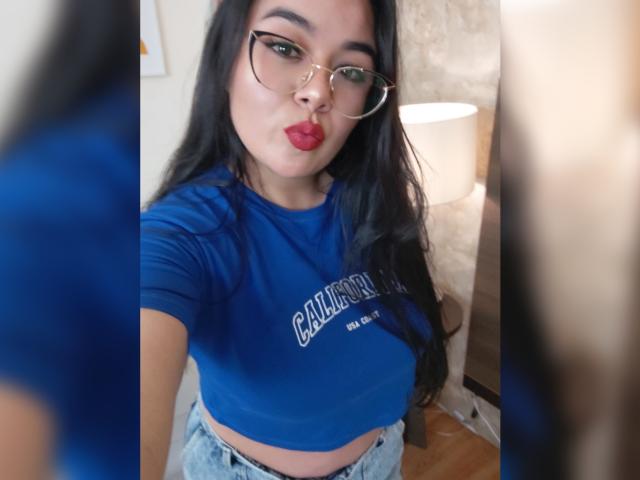 HannahMolly, avec de longs cheveux noirs, portant des lunettes et un débardeur bleu, les lèvres rouges pincées, montrant ses petites seins, dans une pièce avec une lampe allumée..