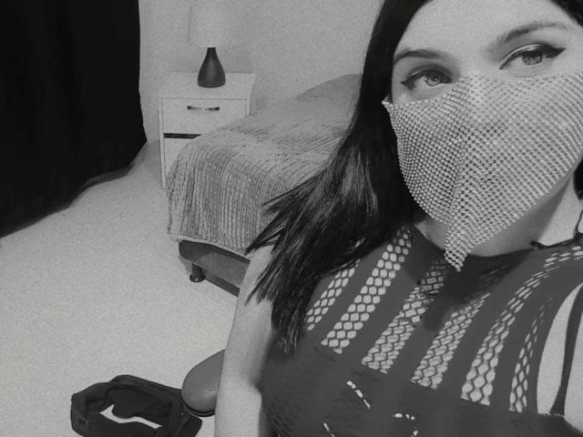 AniisaAli with black mesh mask, wearing a see-through black top, showing her tits, in a grayscale selfie. She has dark hair, light skin, and a small, dark vase.