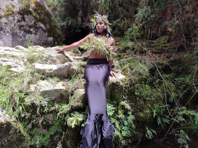 Caylin with black mermaid-style dress, holding green foliage, sitting on moss-covered rocks, forest background, wearing leaf crown, long black tail, slim with visible tits, dark skin.
