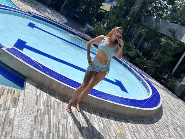 SaarahJones with tan skin, red hair, and tattoos, wearing a blue crop top and mini skirt, standing by a blue-tiled pool on wooden decking, looking up, smiling.