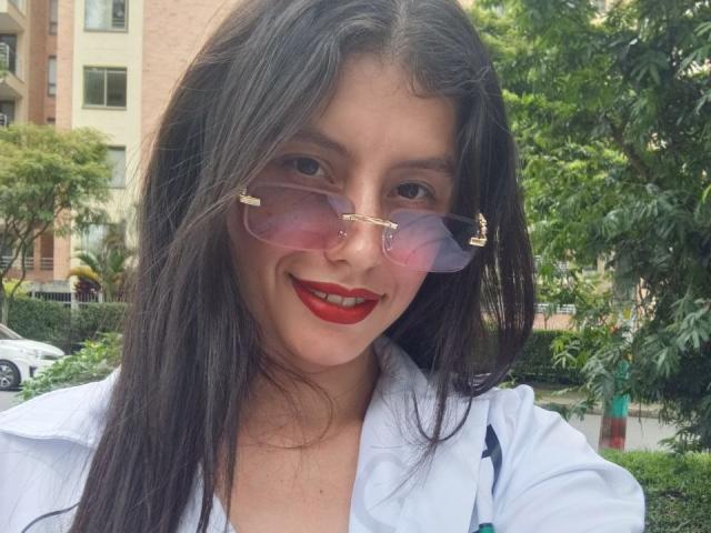 Madisson Fontaine, con capelli neri e lisci, indossa occhiali da sole rosa con dettagli dorati e rossetto rosso, sorride con la sua camicia bianca, in un ambiente esterno soleggiato e verde..