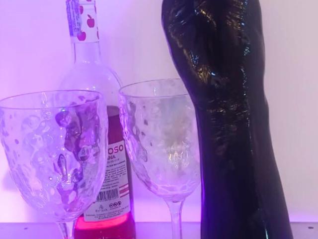 Une bouteille de boisson rose, deux verres à vin vides et renversés avec de l'eau, et un sextoy noir dressé verticalement au premier plan..