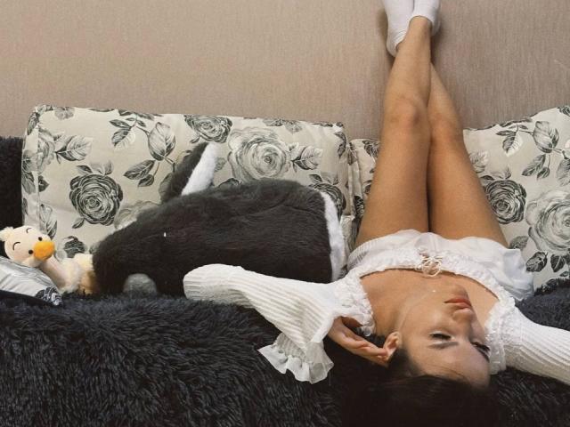 JinnyWalthers lies on a couch with her legs up, wearing a white lace dress, and white socks, with a black stuffed cat and a white duck toy.