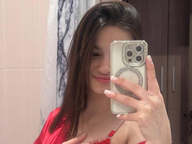 JinnyWalthers with straight brown hair, pink lipstick, and red top, taking a mirror selfie with her right hand, showing off her tits in the reflection.