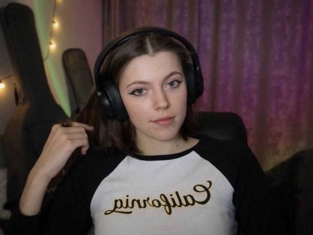 JennellMatacale with black headphones, brown hair, and a white-black raglan shirt, sitting, holding hair, looking at camera, soft smile, slightly upturned nose.