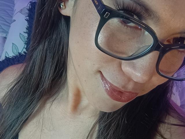 El rostro de Mia Reed, con gafas negras, labios rosados brillantes y cabello castaño, muestra una ligera sonrisa, tiene una pequeña marca marrón en el cuello y pequeños aretes dorados..