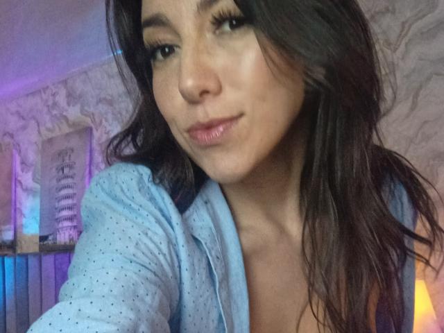 MiaReed with long brown hair, wearing a light blue shirt, has pouty lips and is showing off her tits in a selfie, looking directly at the camera.