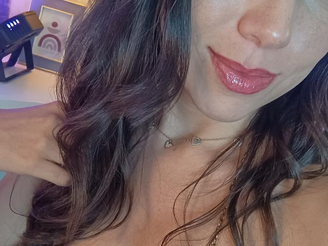 MiaReed's face with glossy red lips, wavy brown hair, and a heart necklace, showing off her tits with hands near her neck in a slightly angled selfie.