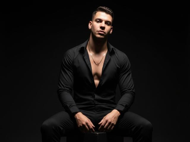 JordanSexxy, sitting with legs spread, wearing an open black shirt, exposing a muscular chest, and a thin necklace, against a dark background. He's looking down with a serious face.