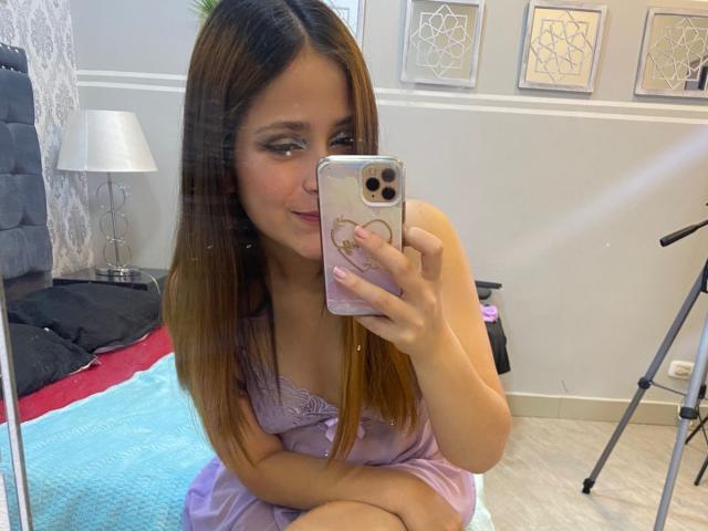 ElisabetLot, con el pelo largo y castaño, vestida con un vestido lila, tomando un selfie con un teléfono dorado, mostrando su cara bonita y sus pequeños senos, sentada en una cama..