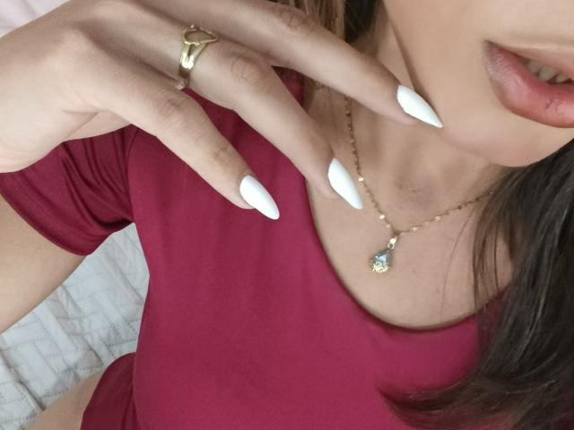 AliceCoolenk's face, showing her pouty lips and white, almond-shaped nails. She's wearing a red shirt, gold necklace with a green gem, and a gold ring.