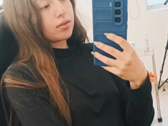 VioletaNova mit langen, braunen Haaren, trägt einen schwarzen, gerippten Rollkragenpullover, hält ein blaues Telefon, macht ein Selfie im Spiegel und zeigt ihre Hand mit ungepinselten Nägeln, wirkt leicht gelangweilt..