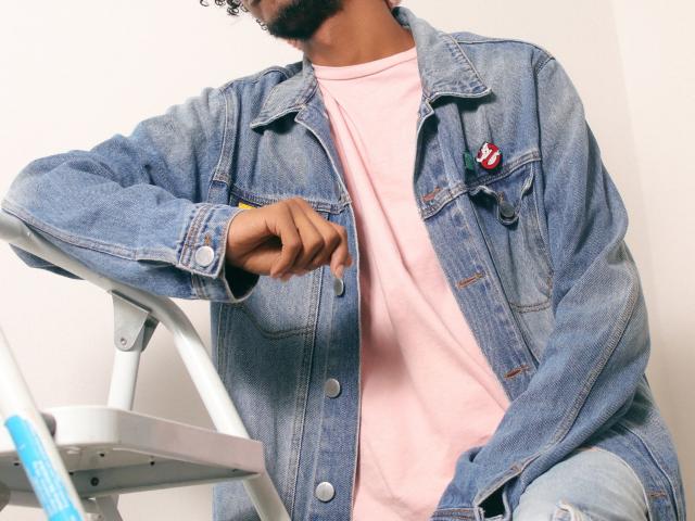 AlexJaccob, que lleva una chaqueta vaquera azul con un parche rojo y blanco, sobre una camiseta rosa, está sentado en una escalera blanca, con el dedo apuntando hacia abajo, y se puede ver su barba rizada..