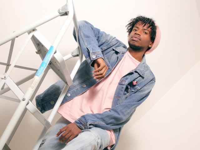 AlexJaccob with curly black hair, wearing a pink beanie, pink t-shirt, blue denim jacket, and light blue jeans, sitting on a metal ladder, looking at the camera.