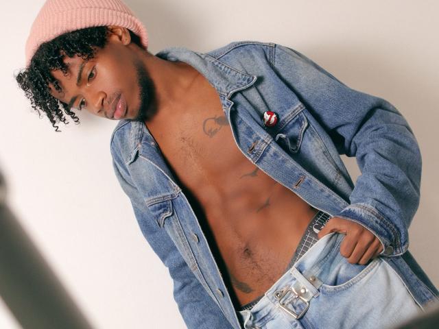 AlexJaccob, con cabello negro rizado, un gorro rosa, una chaqueta vaquera azul, y una camisa desabrochada que deja ver abdominales tonificados y un pequeño tatuaje en el pecho, lleva jeans azul claro y un cinturón negro..