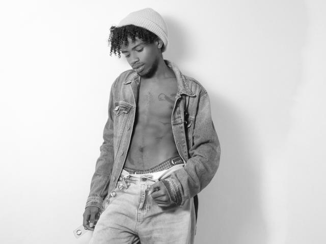 AlexJaccob, shirtless, wearing a beanie, denim jacket, and unbuttoned jeans, showing a tattoo on his chest, with a hand in his jeans, in a black-and-white photo.