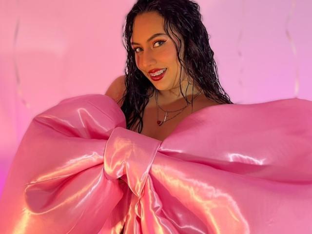 AmyRoss with wet black hair, smiling, covered in a shiny pink satin bow, necklace with heart pendant, pink lighting, looking at camera, pink background with subtle string lights.