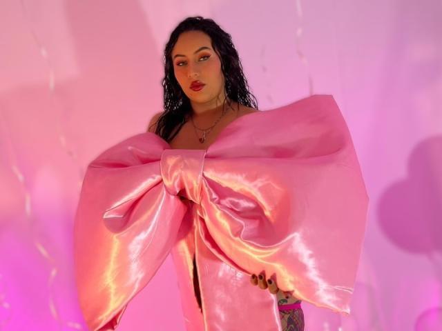 Amy Ross, com cabelo preto molhado, vestindo um robe rosa brilhante de cetim com um grande laço, em pé contra uma parede iluminada em rosa, olhando diretamente para a câmera com batom vermelho..