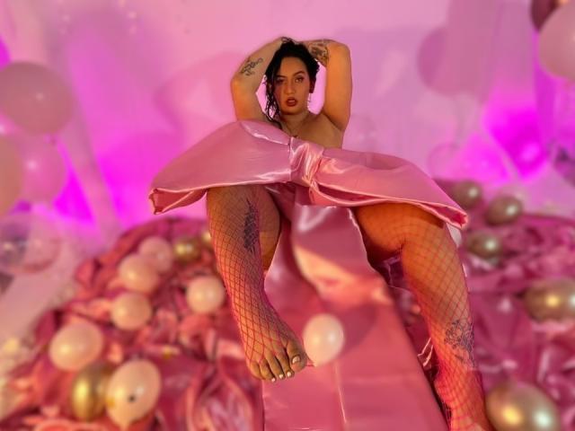 AmyRoss with tattoos, wearing a pink satin dress, fishnet stockings, and no top, legs spread, arms behind head, pink and purple lighting, and gold balloons in the background.