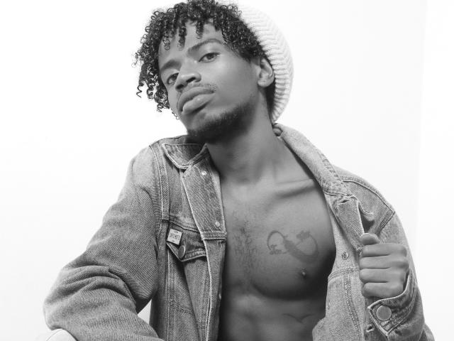 Black-and-white photo of AlexJaccob with curly hair, beanie, and open denim jacket, exposing a tattooed chest, pouty lips, and a confident, sexy expression.